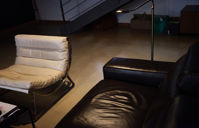 En un sótano iluminado por una lámpara se distinguen un sofá de cuero negro y una silla de metal cubierta con almohadillas blancas. Al fondo varias cajas y mochilas apiladas bajo unas escaleras de metal.