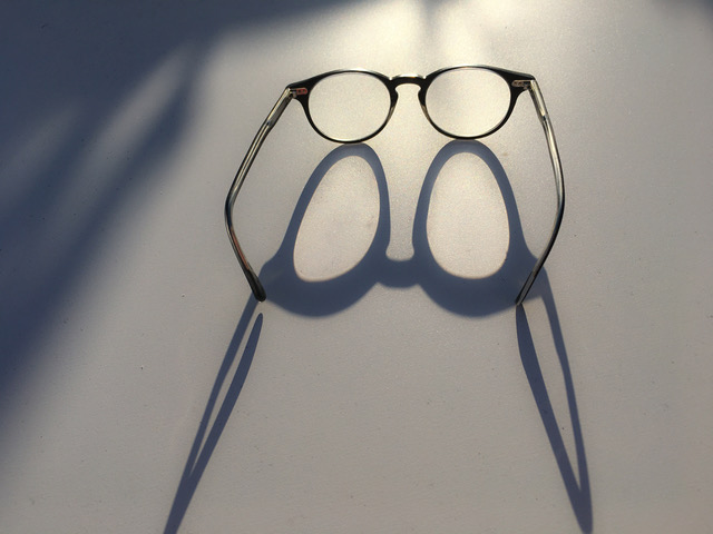 Sobre una mesa gris, vemos unas gafas de lente redonda abiertas mirando al fondo, donde surge un foco de luz que genera una sombra alargada.