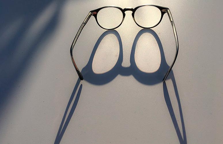 Sobre una mesa gris, vemos unas gafas de lente redonda abiertas mirando al fondo, donde surge un foco de luz que genera una sombra alargada.