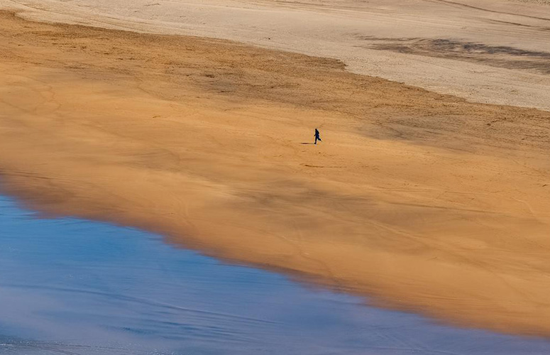 Una playa desierta vista desde el cielo. Se distingue un tercio de arena seca, un tercio de mojada y un tercio de agua que se retrae tras una ola. Sobre la arena mojada se ve la figurita negra de un corredor.