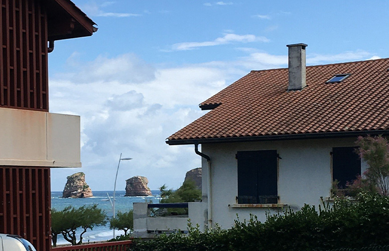 A los lados de la imagen vemos dos casas de pueblo con tejado de teja roja que dejan ver el mar tras ellas. Sobre el mar se alzan dos rocas solitarias en paralelo. Al fondo un cielo azul nuboso y esponjoso.