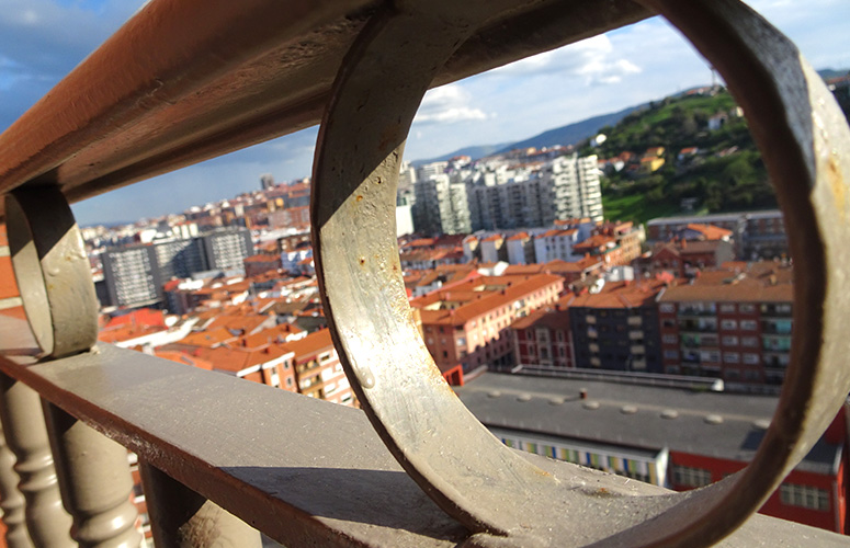 Metalezko baranda baten hutsuneen artetik, hiri baten paisaia ikusten dugu. Antzeko tamaina eta teilatu gorrixkak dituzten eraikin ugari daude, bidegurutze bat osatuz. Eraikuntza zuri eta garaiagoek inguratzen dituzte eta, atzealdean, etxe batzuez zipriztindutako mendixka berde bat dago.