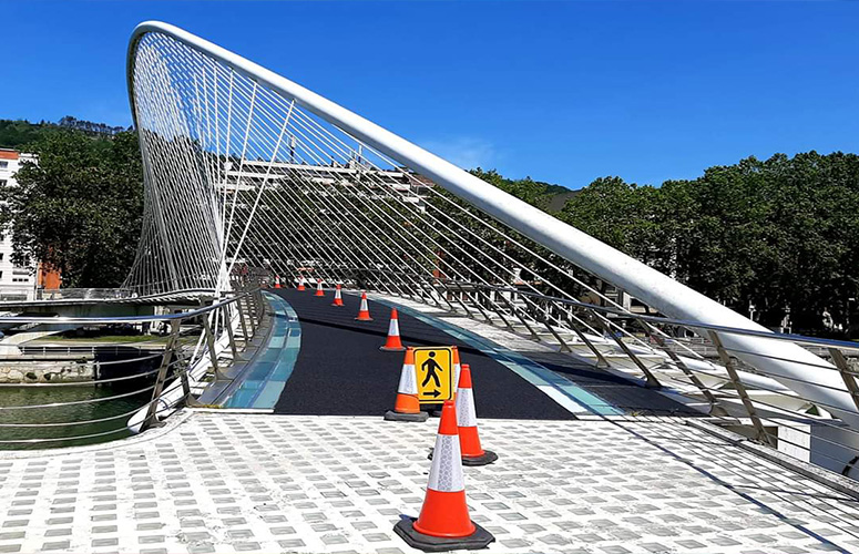Metal zuriko zubi moderno baten bista. Goikaldea, arku lodi batek gurutzatzen du, nondik hari batzuk irteten diren, dena metal zuri berekoak, hesitutako egitura eusten dutenak. Behea kristalez baldosatuta dago eta erdian puntaz punta zeharkatzen duen alfonbra irristagaitza bat jarri da. Bidea trafiko kono gorri-zuriz banatuta dago. Hauen artean seinale horia bat dago jarrita, persona ibiltari baten irudi eta gezi batekin. Azpian, ibai verde batek zeharkatzen du eta, atzealdean, zuhaitz eta eraikin ertain batzuk ikusten dira.