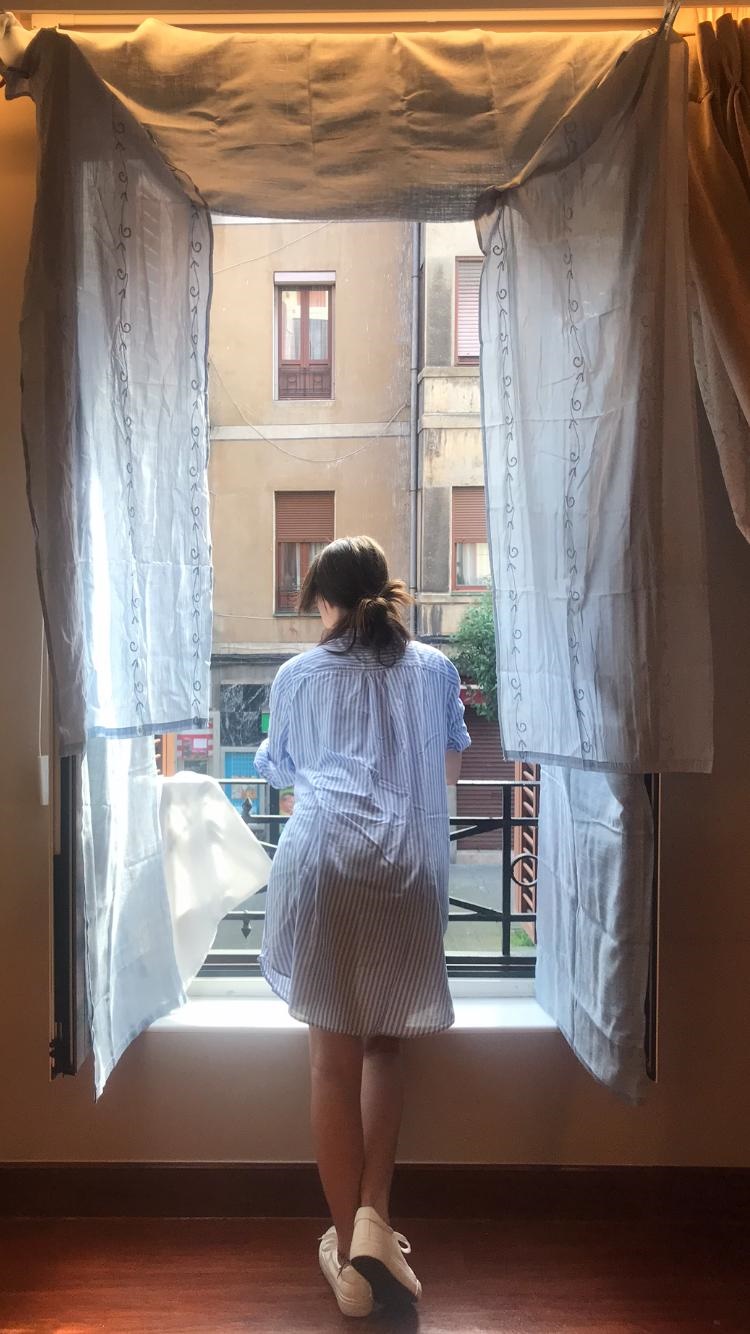 Vemos una imagen pálida, en tonos blancos y luz tenue. En el centro una gran ventana abierta, revestida de cortinas blancas por la que se asoma una chica apoyada en la barandilla de metal, de espaldas mirando hacia el exterior. Viste una bata corta también blanca y lleva el pelo marrón atado en un moño bajo improvisado.