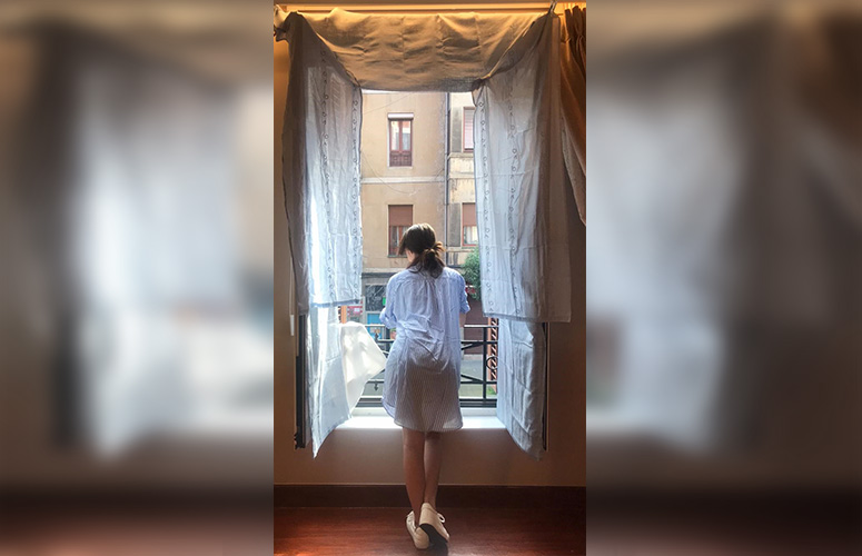 Vemos una imagen pálida, en tonos blancos y luz tenue. En el centro una gran ventana abierta, revestida de cortinas blancas por la que se asoma una chica apoyada en la barandilla de metal, de espaldas mirando hacia el exterior. Viste una bata corta también blanca y lleva el pelo marrón atado en un moño bajo improvisado.