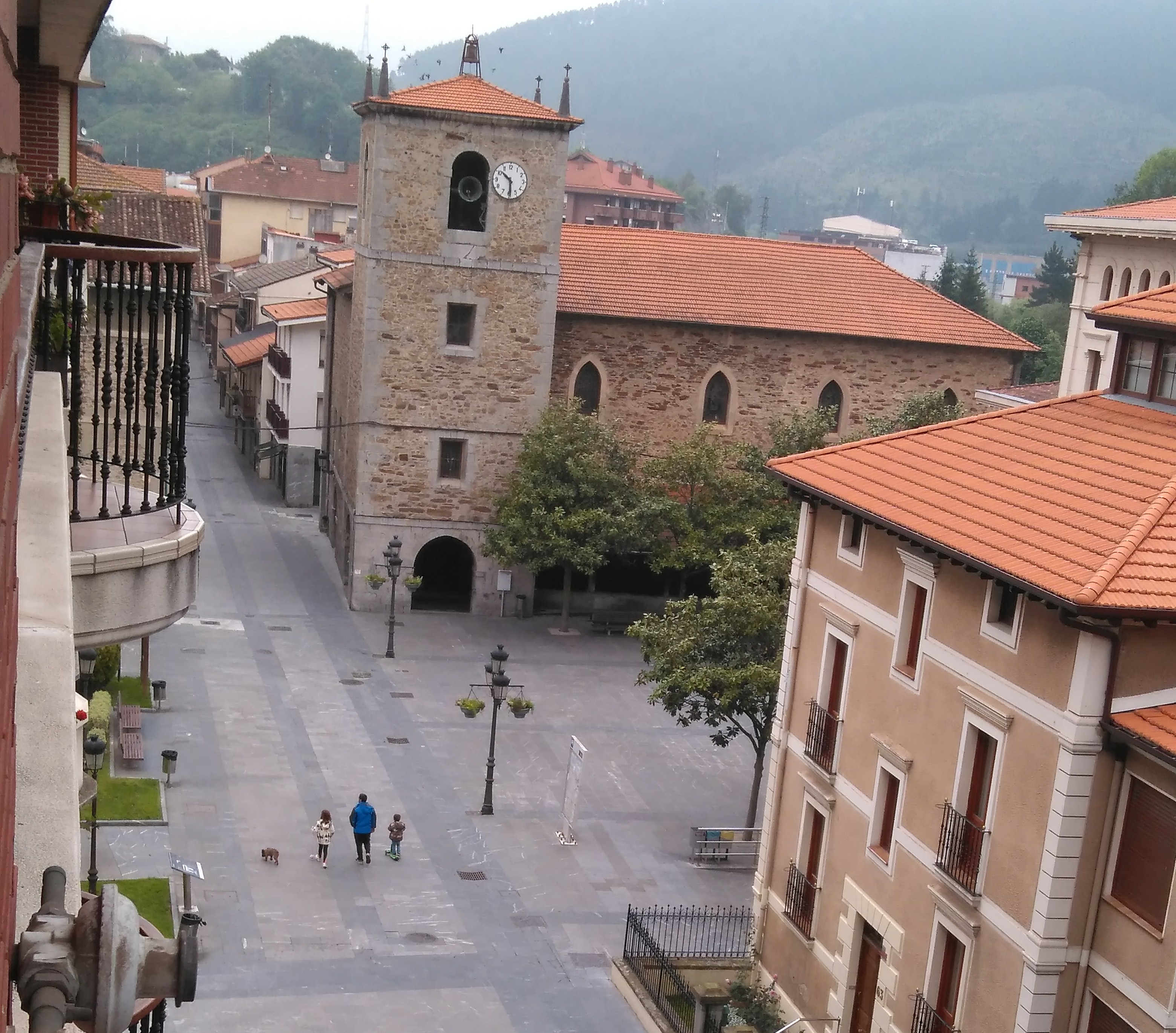 Desde un balcón vemos una calle peatonal que se abre a la plaza de un pueblo con la iglesia de frente rodeada de edificios tradicionales de teja roja. La pequeña iglesia, de piedra y con 4 pequeñas ventanas ojivales en el cuerpo principal. La torre, de 3 pisos, se abre en un pequeño arco desde el que se ve la campana, con un reloj al lado. La calle está desierta salvo por tres personas, un adulto y dos niños, que caminan de espaldas al fotógrafo.