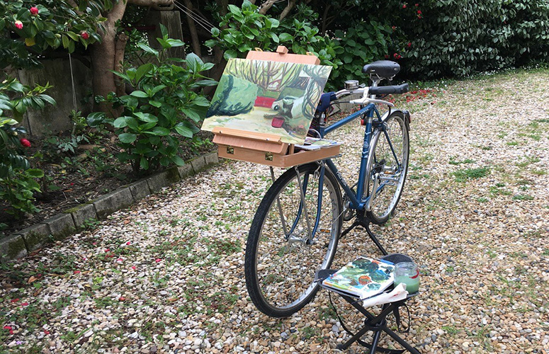 En un jardín de suelo pedregoso parcheado con hierbas y rodeada de vegetación variada, una bicileta azul sirve de soporte improvisado para sostener una caja-caballete en la que descansa un lienzo que muestra varios setos con árboles al fondo y un vehículo blanco. En la esquina derecha una sillita sostiene la paleta manchada de pintura y varios útiles de dibujo más.