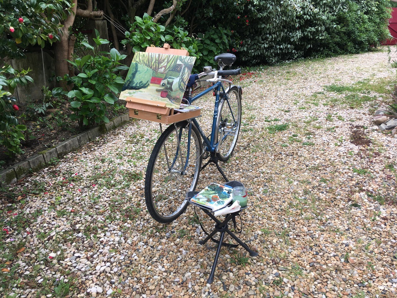 En un jardín de suelo pedregoso parcheado con hierbas y rodeada de vegetación variada, una bicileta azul sirve de soporte improvisado para sostener una caja-caballete en la que descansa un lienzo que muestra varios setos con árboles al fondo y un vehículo blanco. En la esquina derecha una sillita sostiene la paleta manchada de pintura y varios útiles de dibujo más.