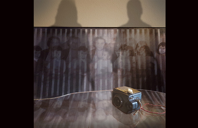 Frente una pared marrón con gotelé, una fotografía antigua partida en rayas muestra a una familia numerosa posando. En primera fila se distinguen tres niños a la derecha, tres niñas a la izquierda y un bebé en el centro sostenido por una mujer. En segunda y tercera línea varios adultos cada cual más desdibujado. Frente al cuadro, una mesa de cristal con una cámara antigua negra de la que pende un cordel rojo.
