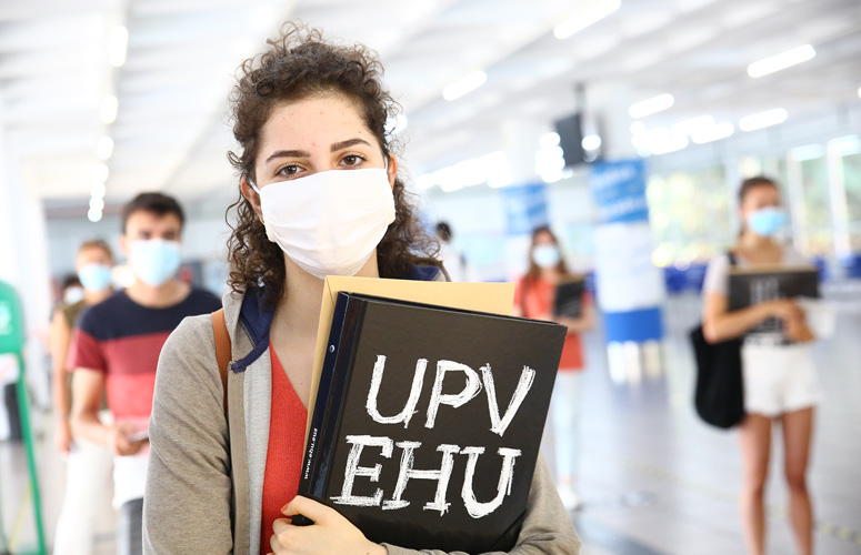 En una amplia sala blanca vemos una hilera de estudiantes con la mascarilla puesta y carpeta de la UPV/EHU en mano. La primera, y única enfocada, es una chica delgada de pelo rizado largo castaño y ojos grandes del mismo color.