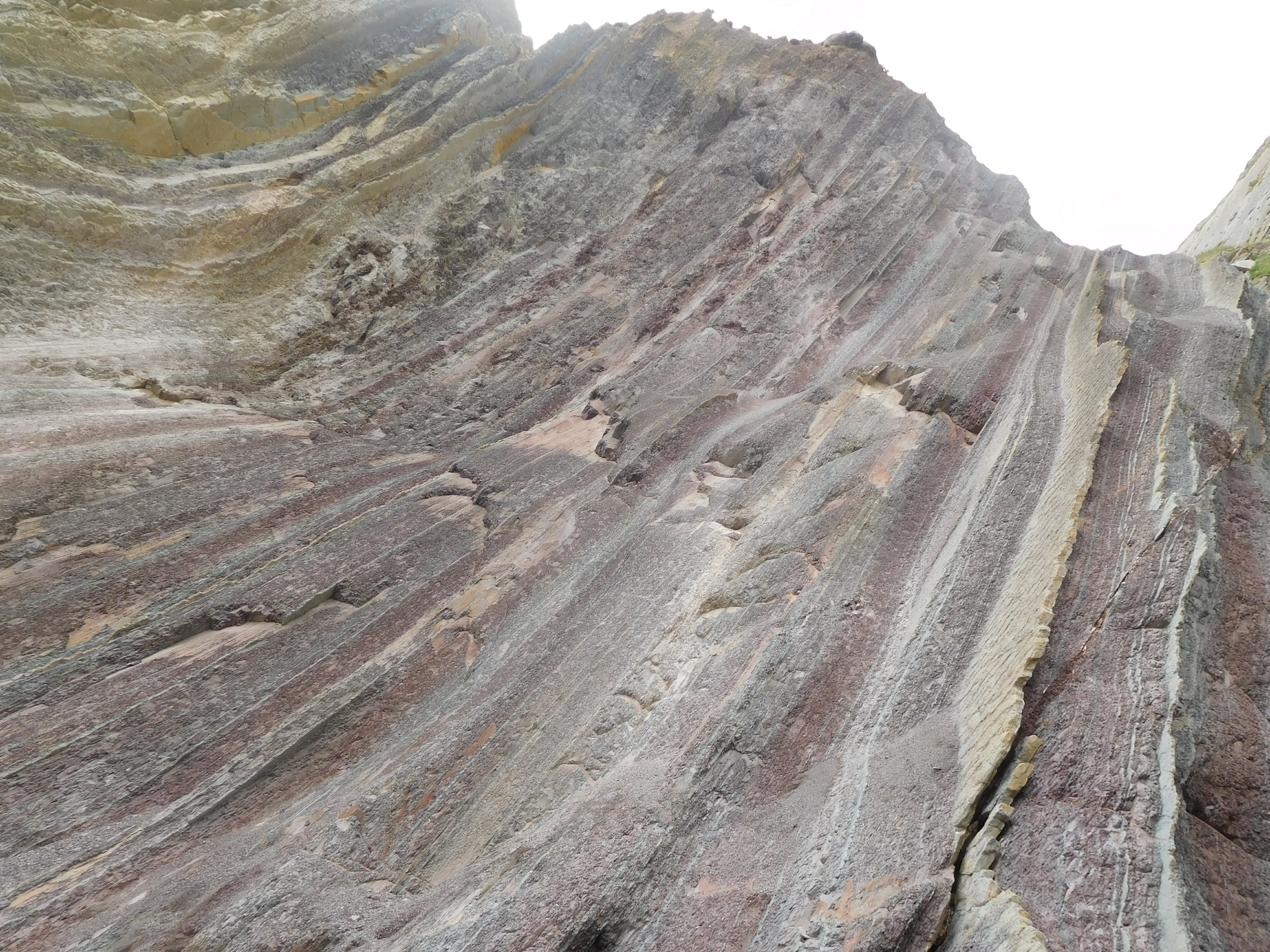 Vemos una formación rocosa ascendiendo como una ola formada por láminas verticales de colores diversos, de naranja a morado y tonos grises. Al fondo se ve un cielo blanco brillante.