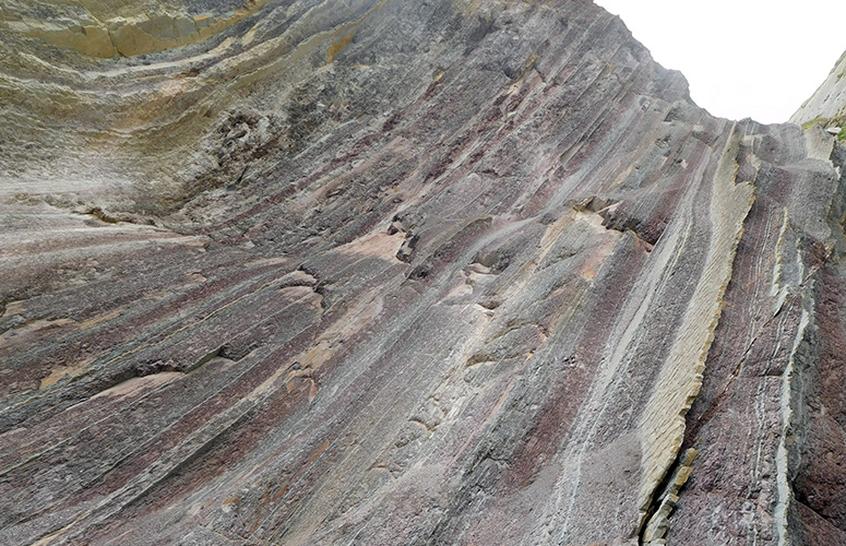 Vemos una formación rocosa ascendiendo como una ola formada por láminas verticales de colores diversos, de naranja a morado y tonos grises. Al fondo se ve un cielo blanco brillante.