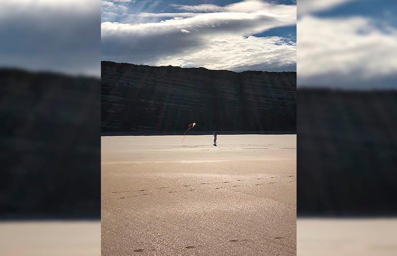 Vemos una playa vacía de arena mojada y lisa sobre la que se distinguen algunas pisadas. En el centro, frente a un gran risco, una niña juega con su cometa. El cielo es nuboso y brillante.