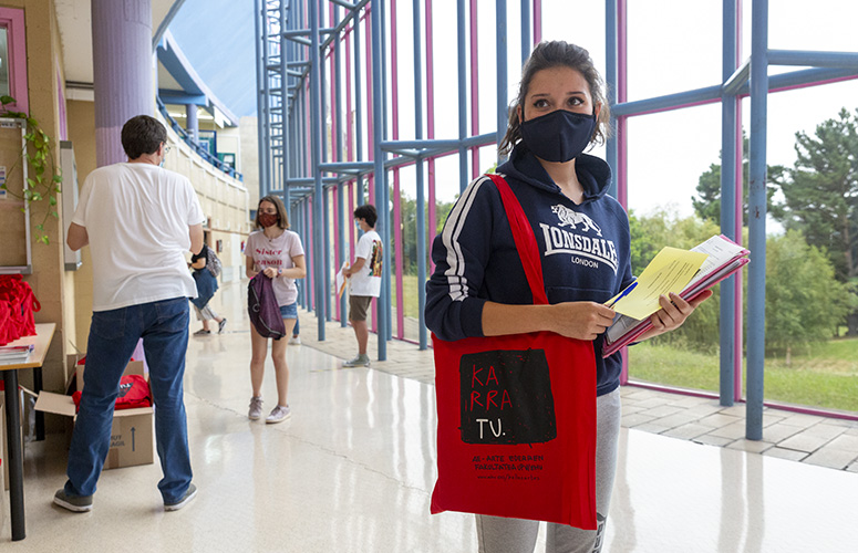 En una amplia sala blanca vemos una hilera de estudiantes con la mascarilla puesta y carpeta de la UPV/EHU en mano. La primera, y única enfocada, es una chica con gafas, delgada de pelo largo castaño y ojos grandes del mismo color.