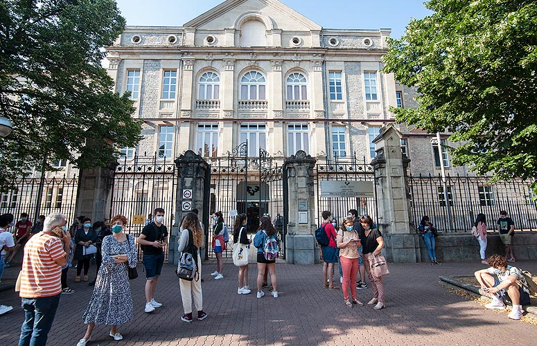 En un aula de biblioteca, se ven varias mesas color madera para cuatro separadas por paneles centrales. En cada una se sienta un alumno con mascarilla y cabeza baja. En las mesas se pueden ver las páginas de los exámenes, gomas de borrar, lápices y bolígrafos.
