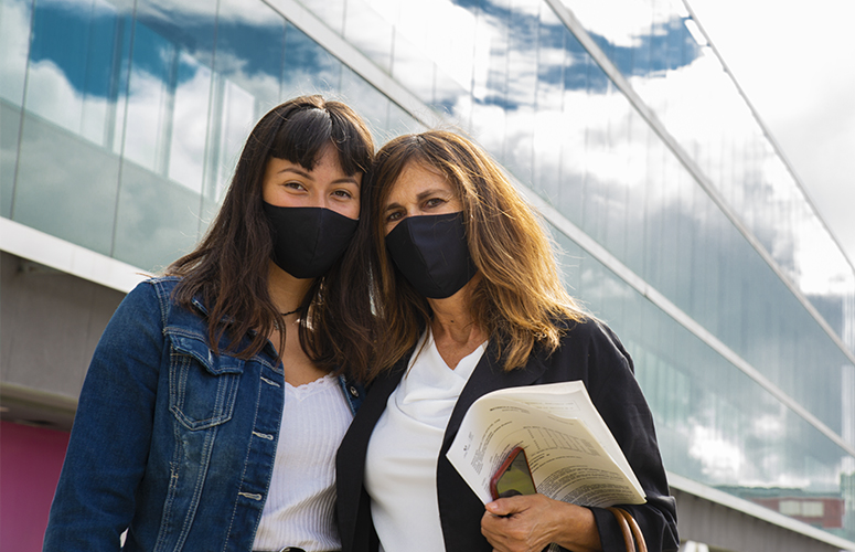 Frente a un edificio acristalado, posan dos mujeres -una hija y una madre- con mascarilla negra. Ambas tienen el pelo marron ondulado y visten blusa blanca bajo una chaqueta, vaquera en el caso de la hija y negra en el de la madre. La madre porta unos papeles y un móvil.